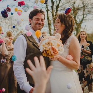 This image shows a bride and groom outside with some other guests standing around them. The guests are throuwing pompom confetti. The couple are in the foreground and the shot is from the knees up. The couple are standing side-by-side with the groom on the left and the bride for the right.The bride has dark hair which she is wearing loose. She's wearing an a-line sleeveless dress with the lace detailing at the shoulders. In her left hand she carries a bouquet of muted pink and white peonies. The groom has dark hair and is wearing a white shirt and a brown tweed waistcoat. The groom has his eyes closed and is smiling the bride is looking at her groom and laughing. In the background there are some trees without leaves. In the foreground there is a hand throwing confetti.