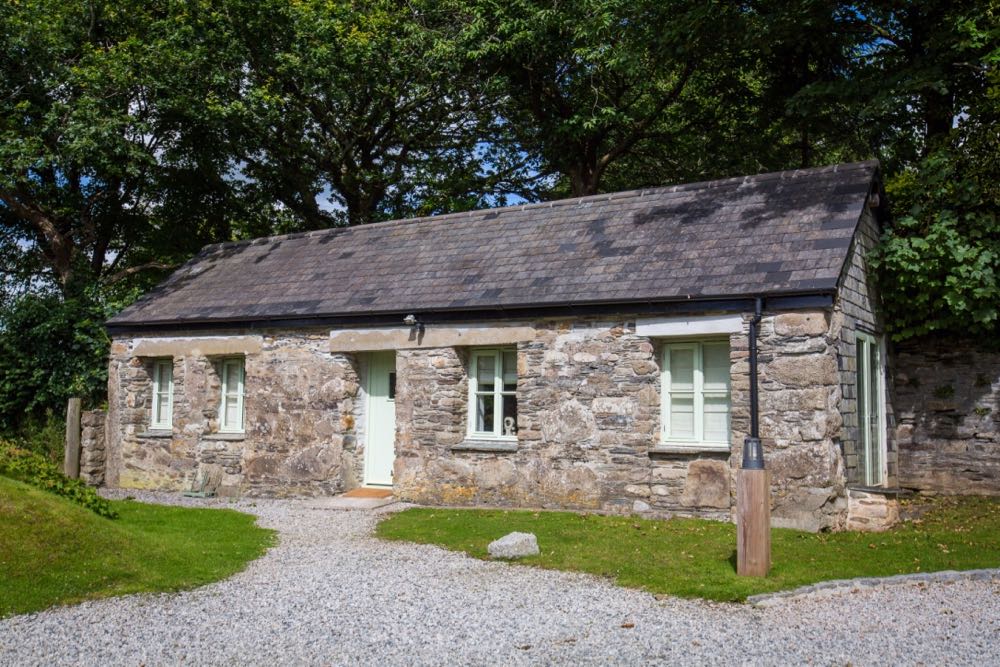 This is a photo of the exterior of cottage number one. Cottage number one sleeps two people. The proerty is single storey. The photo shows the full width of the cottage. In the background you can see trees which are in leaf. In the foreground you can see two patches of grass one on the left and one on the right and in the front there is gravel. The cottage is made of granite stone and is brown and grey in colour. The roof is constructed of grey slate and is pitched with the roof line running from left to right of the photograph along the length of the cottage. In the middle of the front of the cottage there is a wooden door that is painted pale green on either side of the story there are two windows on each side of this door. They are georgian style sash windows and also painted green. On the right hand side of the cottage the gable end has hanging slate on it. There is a French window in this end of the cottage and it is also painted green. In the foreground of the picture there is a light bollard for outside lighting.