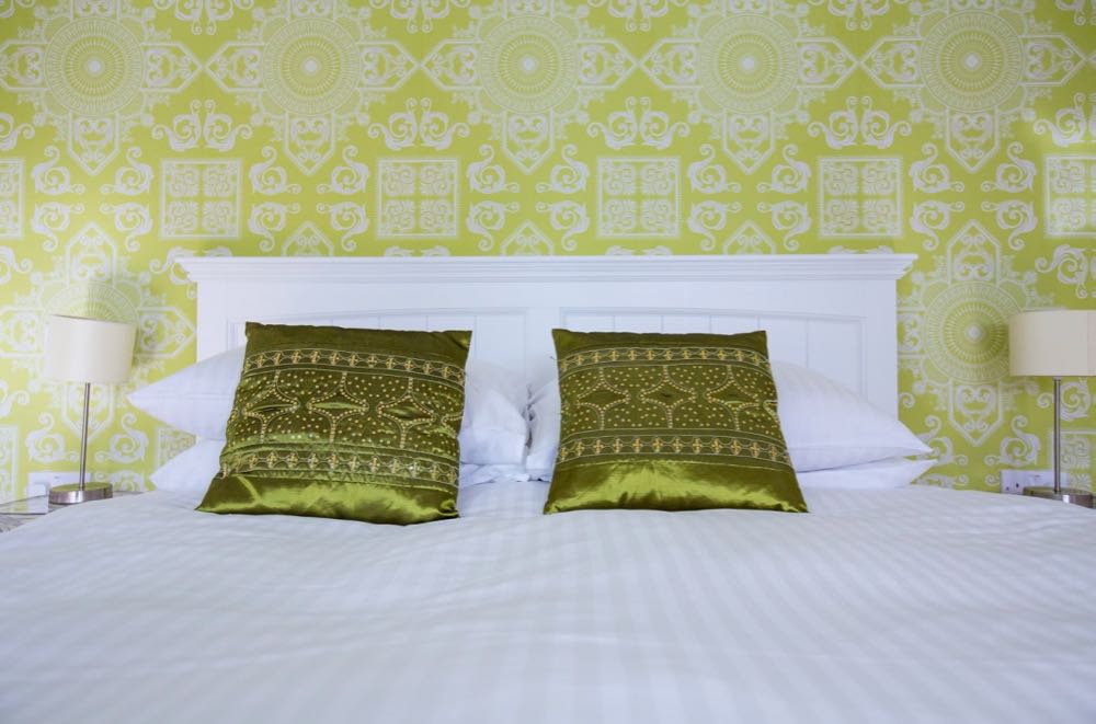 This picture shows a detail from the bedroom in cottage number one which is the small cottage that sleeps two people. The image shows the headboard and top part of a double bed. In the background there is some wallpaper and on each side of the bed there is a bedside lamp. The headboard is wooden and white. There are 2 pillows in white pillow cases on each side of the bed. The wall paper is lime green with an indian inspired white pattern. There are two scatter cushions one on each side. These are dark, lime green and are beaded in an indian pattern. The bedside lights are simple with cream shades and stainless steel stand. The duvet cover is white.