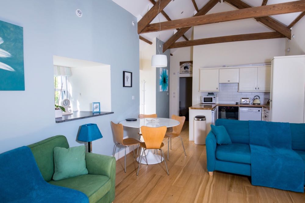 This picture is of the interior of the lounge in cottage number two which is a detached cottage that sleeps four people. The picture shows the lounge / diner area. The photograph is taken looking towards the kitchen area which is furthest from the camera. On the left there is a wall with a window. The window has a striped blind and a slate window sill. On the windowsill there is clock, a potted orchid and a small blue-framed picture. At the back of the room on the left there is a doorway to another room. To the right of this is the kitchen area. The whole room has an oak floor. There are old a-frame beams in the vaulted ceiling and you can see 2 of them in this picture. The wall on the left is painted pale blue the ceiling is painted white and the rest of the walls are painted cream. In the lounge area towards the front of the photograph there is a 2-seater green sofa with a blue throw on the left and a 2-seater blue sofa with a blue throw between the lounge and the kitchen area, facing the camera. There is a table lamp with a blue shade. Between the lounge area and the kitchen on the left there is a dining table. This table is round and white and is on a white pedestal. The chairs are simple dining chairs made of wood with metal legs. There are 4 dining chairs. There is a white pendant light above the dining table light. On the left-hand wall in the lounge there are three pictures with flowers as the subject. The kitchen has got high-level wall units (4 visible on the back wall) and low-level floor units which are cream and the worktop is oak. On top of the worktop you can see a microwave, a toaster and a kettle.