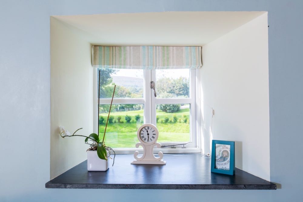 This image shows a detail of a window from cottage number two which sleeps 4 people. The image shows a windowin a wall which is painted blue. The reveals are painted cream and there is a slate windowsill. The window is a Georgian style four pain window. The window has a striped blind which is open. On the windowsill there is a potted orchid, a clock and a small blue frame with a paisley print in it. Outside the window you can see a distant hill, a lawn and a small box hedge.