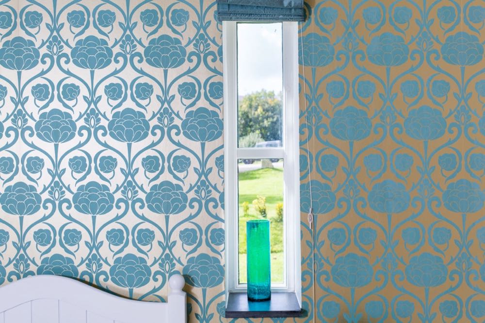 This image shows a detail of a window from cottage number two which sleeps 4 people. The image shows a window in a wall which has pale gold metallic wallpaper with a teal floral design. It is a long narrow window. There is a slate windowsill. The window has 2 panes. The window has a blue blind which is open. On the windowsill there is a teal, glass vase. Outside the window you can see some trees, a lawn and a small box hedge.