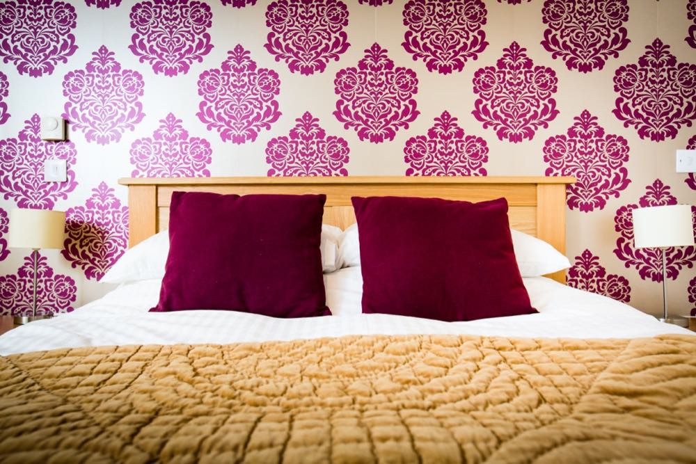 This is a detail shot of one of the double beds in cottage number five. Cottage number five sleeps six people. In this image you can see the wallpaper and the top of the bed. The wallpaper is gold with a red flock pattern. The pattern is quite bold. The bed frame is oak. The duvet cover and the pillows are white linen. There are two red cushions, one on each set of pillows. At the bottom of the bed there is a gold folded throw. On each side of the bed there is a bedside lamp which has a stainless steel base and a cream shade.