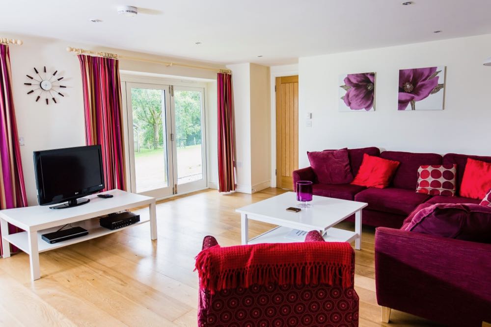 This image is of the lounge area in cottage number five. Cottage number five is a three bedroomed cottage sleeping six people. The room has oak floors. All the walls are painted cream and the ceiling is painted white. There are spotlights in the ceiling. There are two window to the left of the camerabut only one window can be seen fully. This is a bi-fold door which is full height, floor to ceiling. Out of this window you can see trees that are in leaf. The curtains are striped in colours of red brown and gold. There is a 