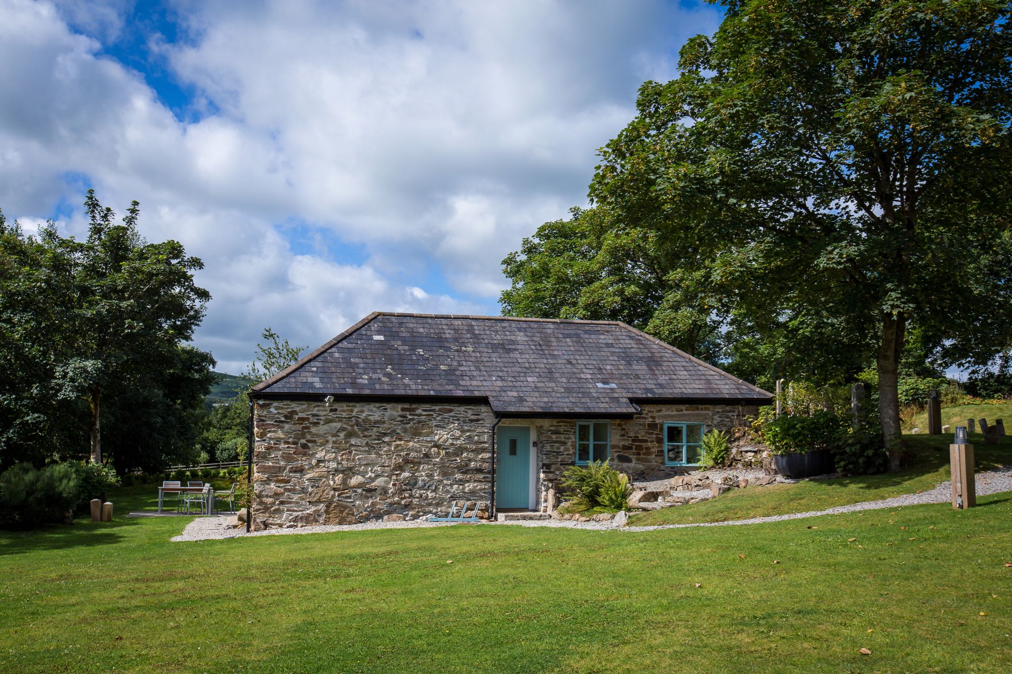 This picture shows cottage number two which is a detached cottage that sleeps 4 people. The cottage is in the middle of the image and there are lawns in front and a blue cloudy sky above. There are trees in leaf to the left and to the right. The cottage is built of local granite stone which is brown and grey in colour. It has a grey slate roof the main roof line runs from left to right along the length of the photograph. In the middle of the building there is the front doorthat is painted pale blue. On the right of the door there are 2 georgian style sash window also painted blue.