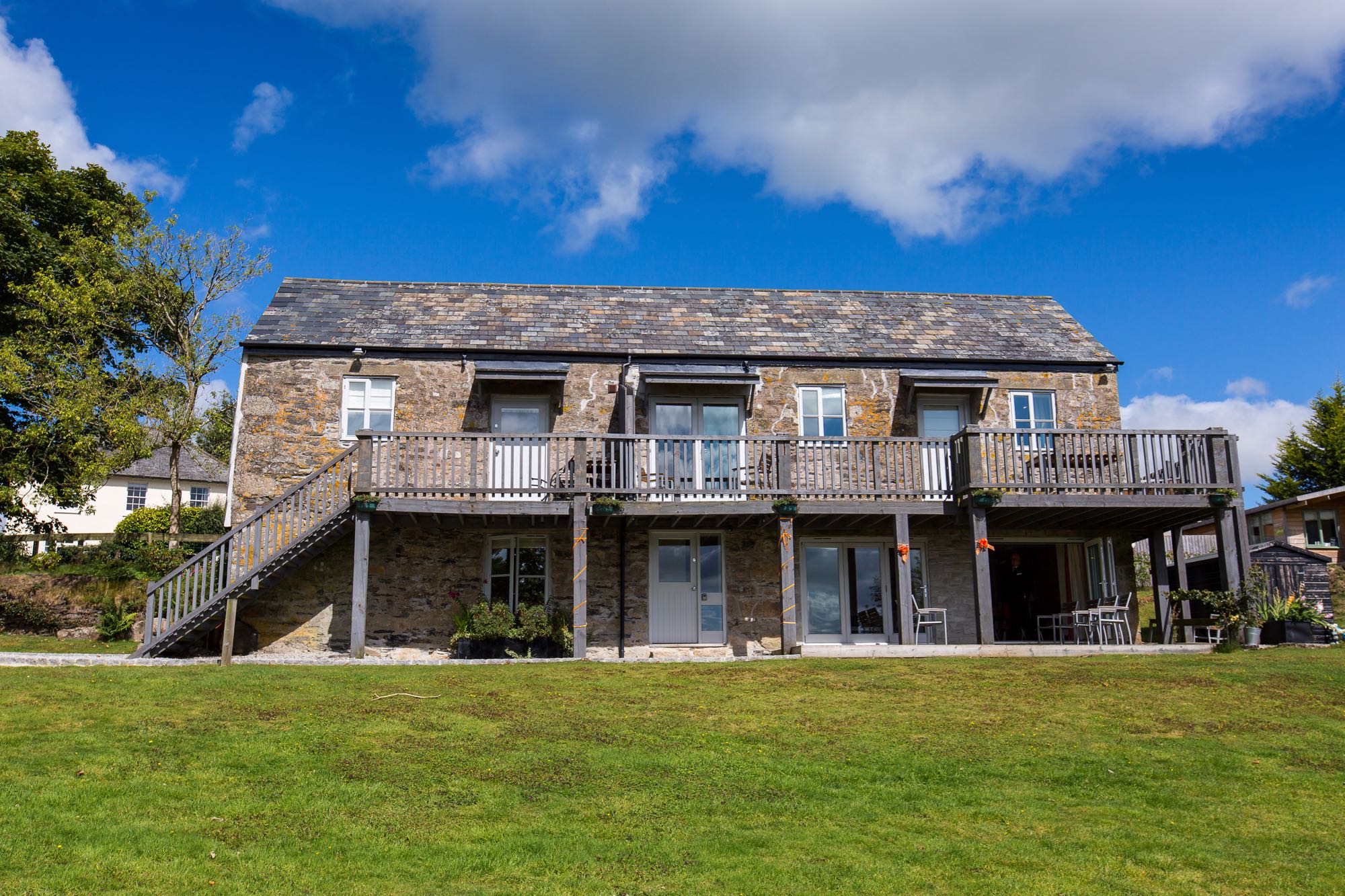 This is an exterior shot of a large barn that is split into three cottages namely cottage three, cottage four and cottage five. The barn has two storeys. Cottage three and cottage four are upstairs, 3 on the left, 4 on the right. Cottage five is on the whole of the ground floor. In the image the large barn is seen from the front. The barn is made of local grantie cob that is greay and brown in colour. All the windows and doors are painted pale gray. Along the top floor from left to right there are: a 4 pane casement window, a half glazed door, a set of french doors, a half glazed door and a 4 paned casement window. On the ground floor from left to right there is a 4 paned casement window, a half glazed door with light oanel to the right, a set of bifold dorrs (closed) and a set of bifold dorrs (open). To the left of the building there is a set of grey painted wooden steps leading upwards to the second floor of the building. There is a balcony along the full length of the second floor. Teh balcony wraps around on the right hand side. The balcony is painted grey and is made of wood. The balcony is supported on wooden posts from the ground. The area immediately around the cottage is a gravel path and at the far end of the front of the building (on the gorund floor) you can see a decking area with some outside chairs and a table. All around the gravel path is lawn and to the left and to the right are trees in the leaf. The sky is blue with some clouds. to the left of the picture you can see the white farmhouse in the background.