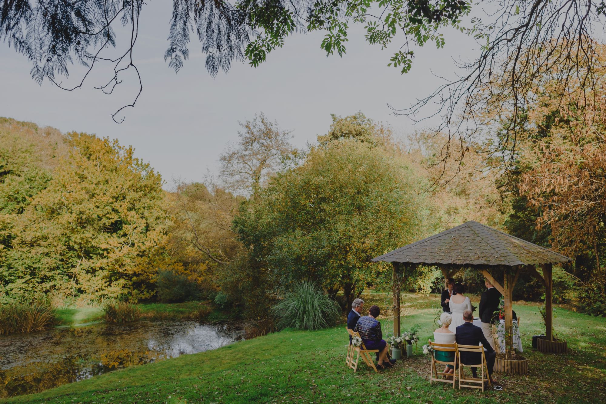 This photo is taken at the lake arbour. The Lake Arbour is in the middle of the photograph. There is a couple standing under the Lake Arbour. They are exchanging their vows. Also under the Lake Arbour are the registrar and her assistant. There is a table covered with a white cloth. The photo is taken from the left of the arbour and the arbour fills the bottom quarter of the shot. Youc an see the lake and lots of autumn trees in the background. The Lake Arbour is a four posted wooden structure supporting a slate roof which is gray. The structure is made of oak. The whole of the ground around the area is lawn. There are climbing shrubs around each of the four posts of the arbour and the front of the arbour is decorated with some hanging folliage. The couple are standing with the bride on the left and the groom on the right. They are facing the registrars with their backs to their guests, and their backs to the camera. Ther are some vases of flowers with white flowers at the base of the front, left-hand arbour post. The bride is wearing a knee-length, full-skirted, a-line dress. She has dark hair which she is wearing up. The groom is wearing chinos and a dark jacket. He has light brown hair. There are 4 guests seated on wooden chairs facing the couple with their backs to the camera. There are 