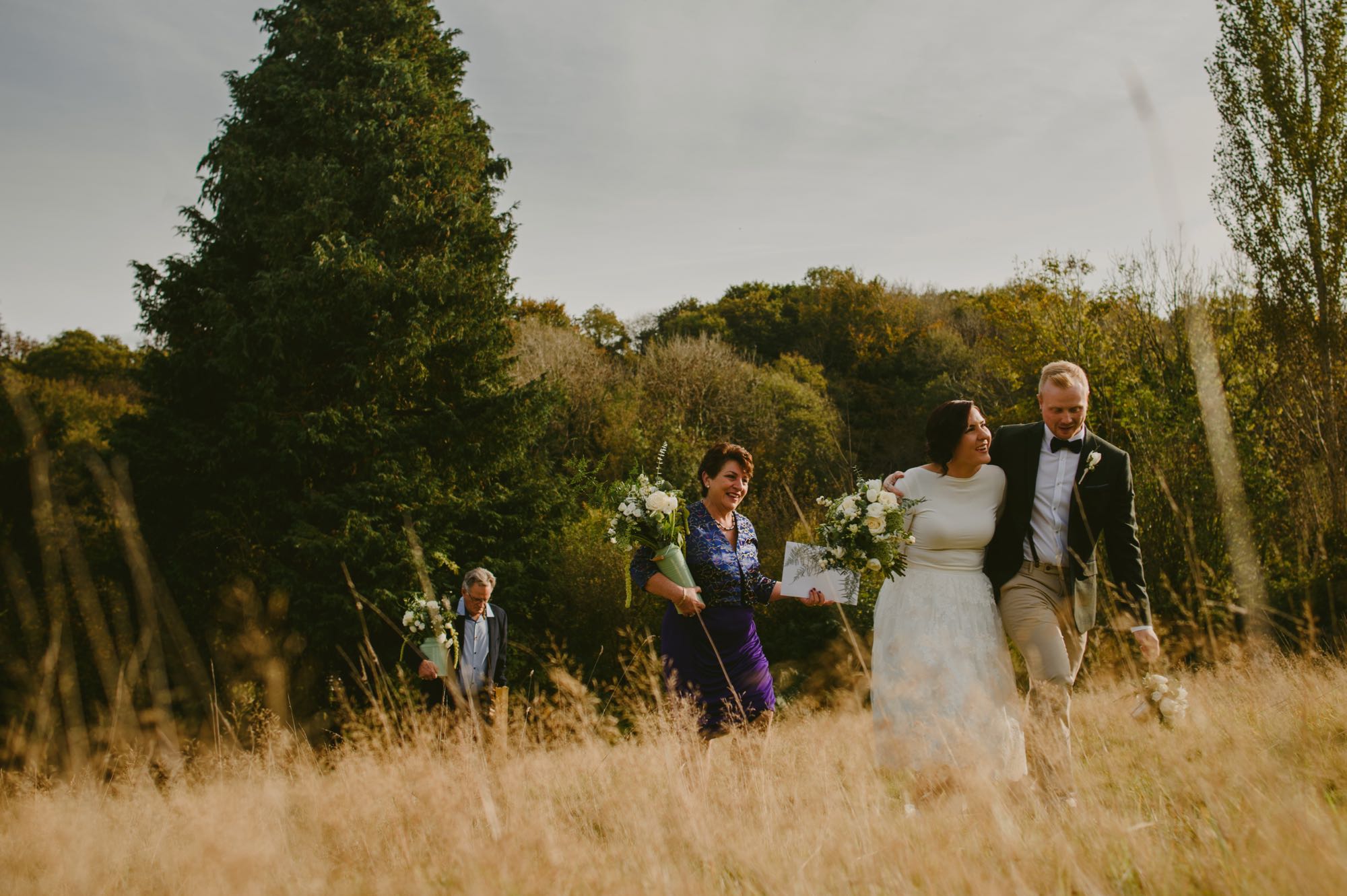 This photo is taken in the meadow with a wedding party of 4 people making their way up a slope from the Lake Arbour. The grass in the meadow is long, parched and golden. There are many trees in the backgorund and they are showing their autumn colours. The couple are in out front walking. The groom is on the left and the bride is on the right. The groom has his arm around his bride's shoulders. They are facing the camera. The bride's bouquet of white flowers and green foliage is in her right hand. The bride is wearing a knee-length, full-skirted, a-line dress. She has dark hair which she is wearing up. The groom is wearing chinos, brown brogues, a dark jacket a pale blue shirt and a dark bow-tie. He has light brown hair. Following behind the couple are the bride's parents. Her mum is wearing a purple pencil skirt with a v-necked silk blouse. Her dad is behind her mum ans is wearing a dark jacket and open necked blue shirt. Both are carrying a large vase of white flowers with green foliage.
