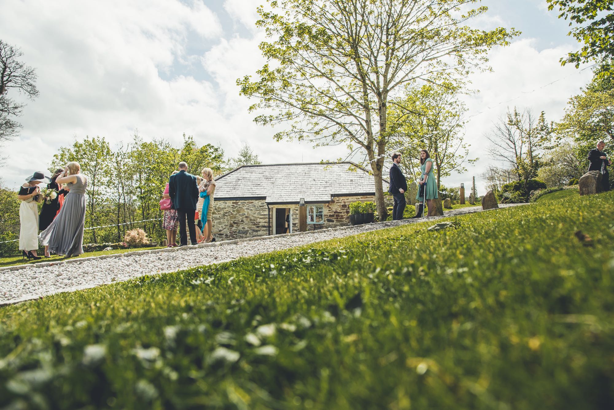 This is an image of some guests relaxing in the grounds. In the middle of the picture you can see cottage number two which is a sleeps two cottage. This cottage is detached and is made of local granite stone which is gray and brown. It has a gray slate roof. There is a door in the middle of the cottage and the door is open. There is a casement window to the right hand side of the door. In the foreground of the picture there is grass and diagonally across the picture there is a gravel pathway sloping upwards. The sky is blue but cloudy. It is a spring day and the trees are partially in leaf. There are trees to the left and to the right of the cottage. On the left of the picture at a distance of about 20 meters from the camera are 3 guests standing in a group. In the middle of the picture, also about 20 meters from the camera, is another group of 4 guests. Towards the right of the picture also at about 20 meters from the camera is another group of 3 guests.