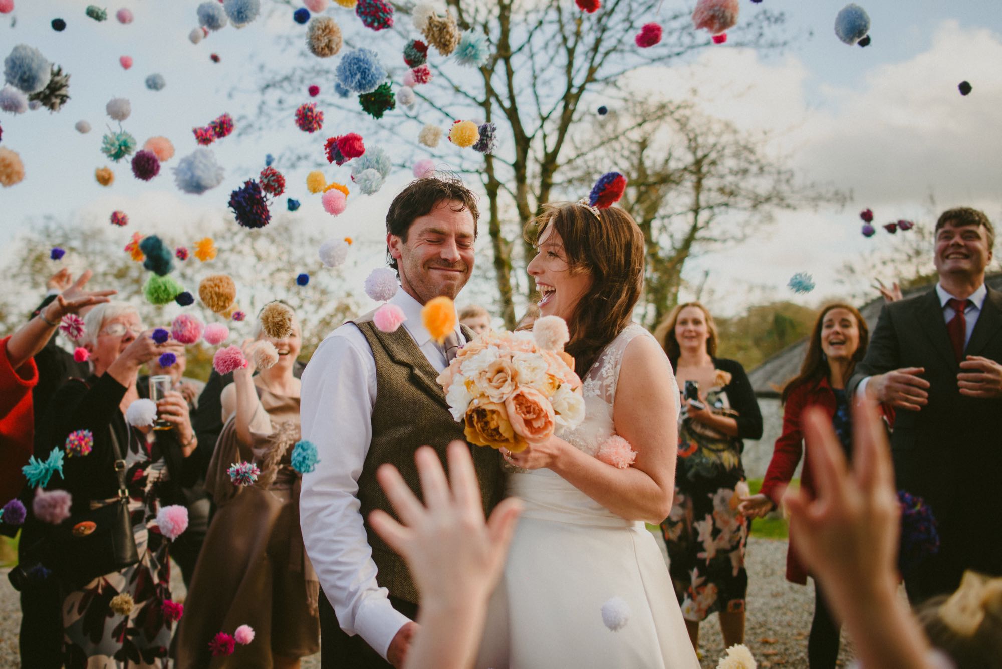 This image shows a bride and groom outside with some other guests standing around them. The guests are throuwing pompom confetti. The couple are in the foreground and the shot is from the knees up. The couple are standing side-by-side with the groom on the left and the bride for the right.The bride has dark hair which she is wearing loose. She's wearing an a-line sleeveless dress with the lace detailing at the shoulders. In her left hand she carries a bouquet of muted pink and white peonies. The groom has dark hair and is wearing a white shirt and a brown tweed waistcoat. The groom has his eyes closed and is smiling the bride is looking at her groom and laughing. In the background there are some trees without leaves. In the foreground there is a hand throwing confetti.