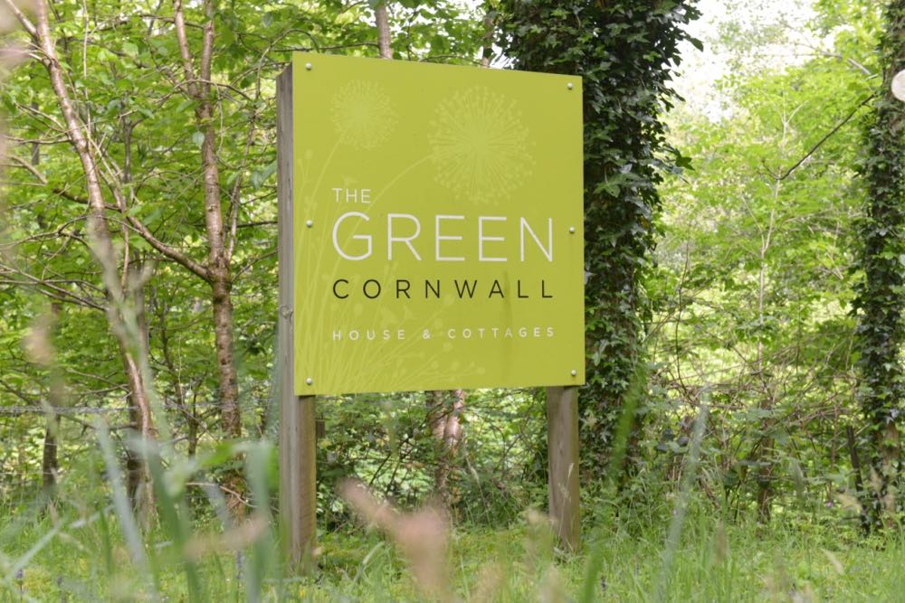 This image shows the sign at the entrance to The Green. The sign is in the middle of the photograph. The sign is square and is lime green in colour. There is a pale dandelion clock motif in the background. There are the words in "The Green House and Cottages" written in white font. The word "Cornwall" is written in black font. This sign is mounted on two wooden posts. The ground around the sign is grass. And there are many trees and shrubs in the background.
