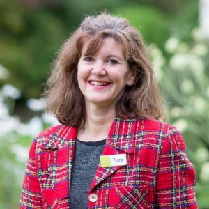 This is a picture of Fiona. Fiona is an event manager and sales manager. This is a head and shoulders shot of Fiona standing outside in front of some trees. She is wearing a red tartan jacket and a grey top. She is wearing a name badge. Fiona has shoulder-length mid-brown hair and she is smiling.