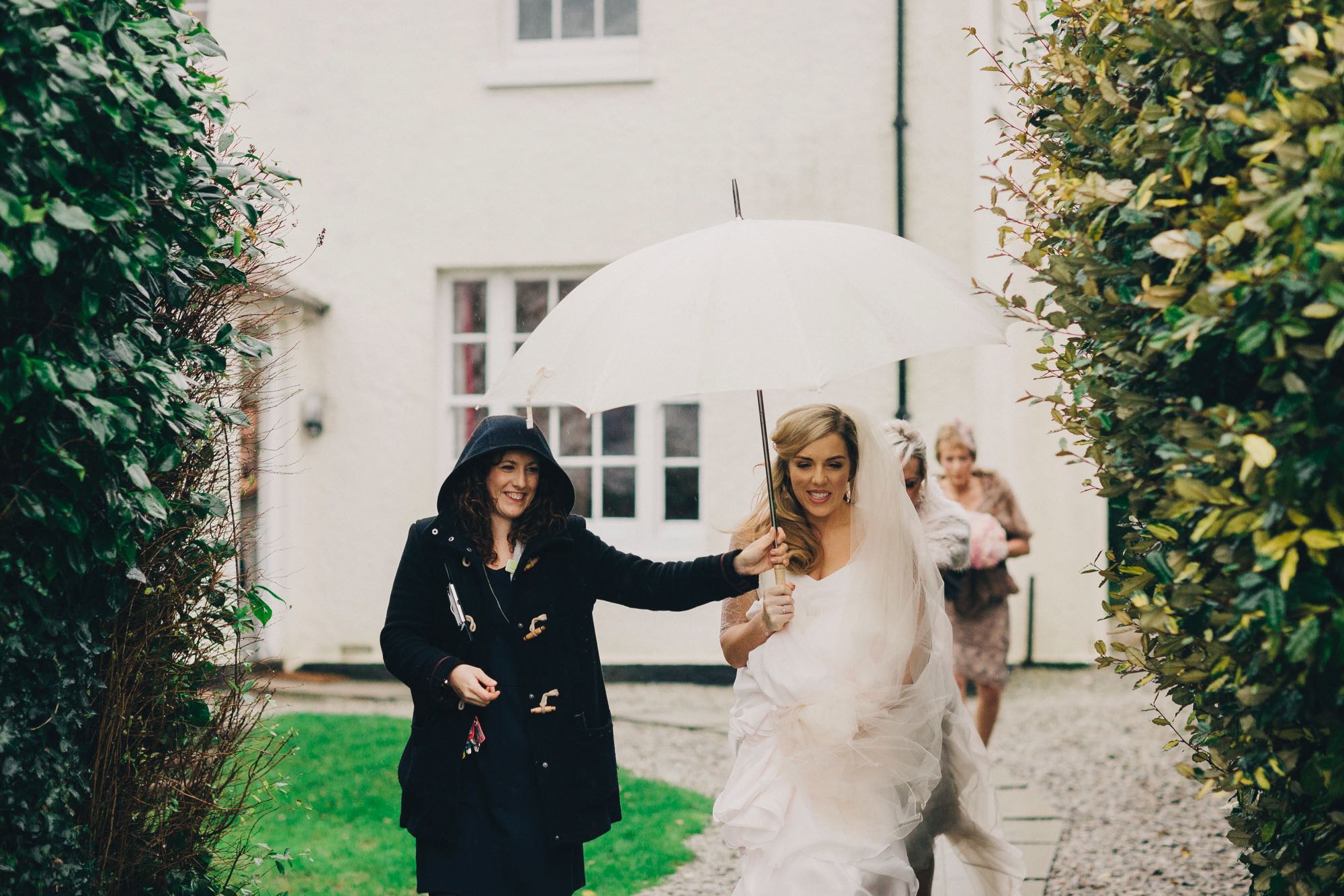 This image shows a member of the Green team staff holding an umbrella over a bride's head as she makes her way towards her ceremony. The picture is taken in front of the farmhouse. There is a bush on either side of the image framing shot. You can see the corner of the house in the background. The farmhouse is white and has Georgian style windows. The member of staff is on the left and she's wearing a blue duffle coat with the hood up as it is raining. She is holding a white umbrella above the bride's head. Both of them are smiling. The bride has dark blond hair that she is wearing loose. She is wearing a short sleeved white gown and a veil. The dress is long and layered and she is holding it up so she can walk. In the background, walking you can see two ladies who are part of the bridal party.