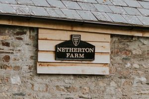 This is an image of the old Duchy of Cornwall farm sign. This sign is made of cast iron which is painted black. The words "Netherton Farm" are spelled-out in capitals in gold lettering. Above the writing is the coat of arms of the Duchy of Cornwall. This is a shield in gold with five rows of dots, one under the other. The first row has five dots, the next row has four dots, the next row has three dots, the next row has two dots and the final row has one dot. The whole of the sign is mounted on 4 wooden boards. The wooden boards are attached to a Cornish granite, cob wall which is brown and gray in colour. This wall forms part of a building and at the top section of the photo part of the roof and guttering can be seen. The roof is made of grey slate. And the guttering is black.