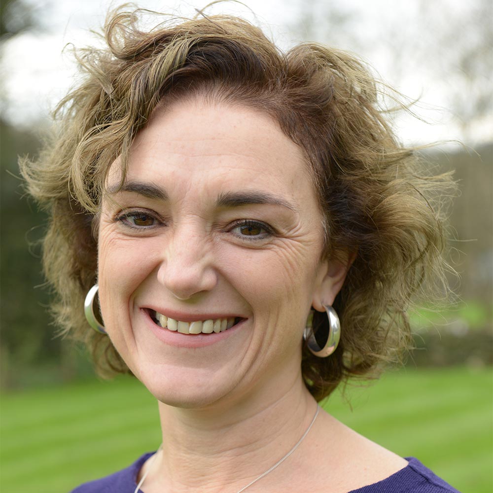 This is a head shot of Moira our sales and marketing manager. Moira is wearing a purple top. She has a silver chain around her neck and large silve, hooped earrings. She has short, dark, curly hair. In the background you can see grass and trees. She is smiling.