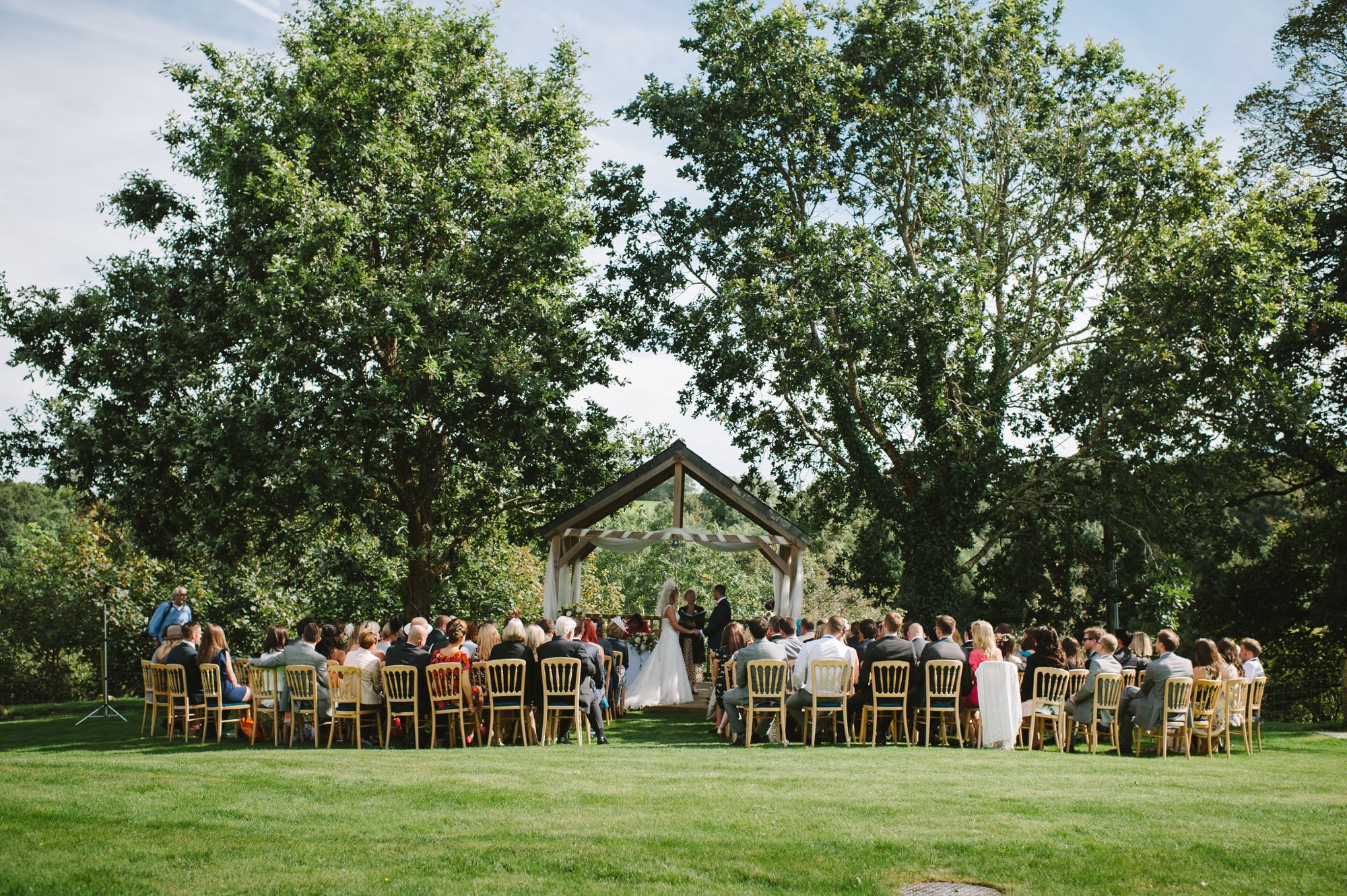 A couple exchanging vows under the Oak Arbour. The arbour is a wooden structure with four corner posts supporting a pitched roof. The arbour has a large Oak Tree on each side. The trees are in full leaf. This photo is taken from a bit of a distance and the whole of the Oak Trees are in the frame. The couple are under the arbour with the registrar and there is a clothed table (white) also under the arbour. The arbour is decorated with a some white voile. The couple are in the middle of the shot but are small compared to the canopy of the trees. They are holding hands and facing each other. The groom is on the right, the bride on the left. It is a summers day and the trees are in full leaf. The bride is wearing a white, full length, sleeveless, full-skirted dress. She has blond hair she is wearing down. The groom is wearing a dark suit. The couple are holding hands and looking at each other. You can see there guests sitting in front of the arbour in two columns of rows of seats.. The guests are looking at the couple towards the arbour and have their backs to the camera. You can see the registrar standing by a clothed table under the arbour with the couple.