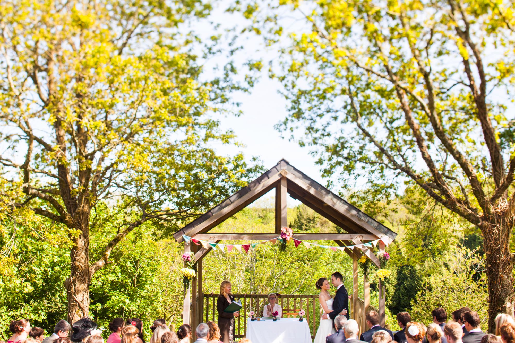 A couple exchanging vows under the Oak Arbour. The arbour is a wooden structure with four corner posts supporting a pitched roof. The arbour has a large Oak Tree on each side. The trees are in partial leaf. This photo is taken from a bit of a distance and most of the Oak Trees are in the frame. The couple are under the arbour with the registrar and there is a clothed table (white) also under the arbour. The arbour is decorated with bunting. The arbour is in the middle of the shot and the couple are standing in the right of the arbour. They are holding embracing and facing each other. The groom is on the right, the bride on the left. It is a spring day. The bride is wearing a white, full length, strapless, a-line dress. She has dark hair she is wearing up. The groom is wearing a dark suit. You can see top of their guests heads as they sit in front of the arbour in two columns of rows of seats.. The guests are looking at the couple towards the arbour and have their backs to the camera. You can seee the registrar holding a book in the right hand corner of the arbour and there is a white, clothed table under the arbour.