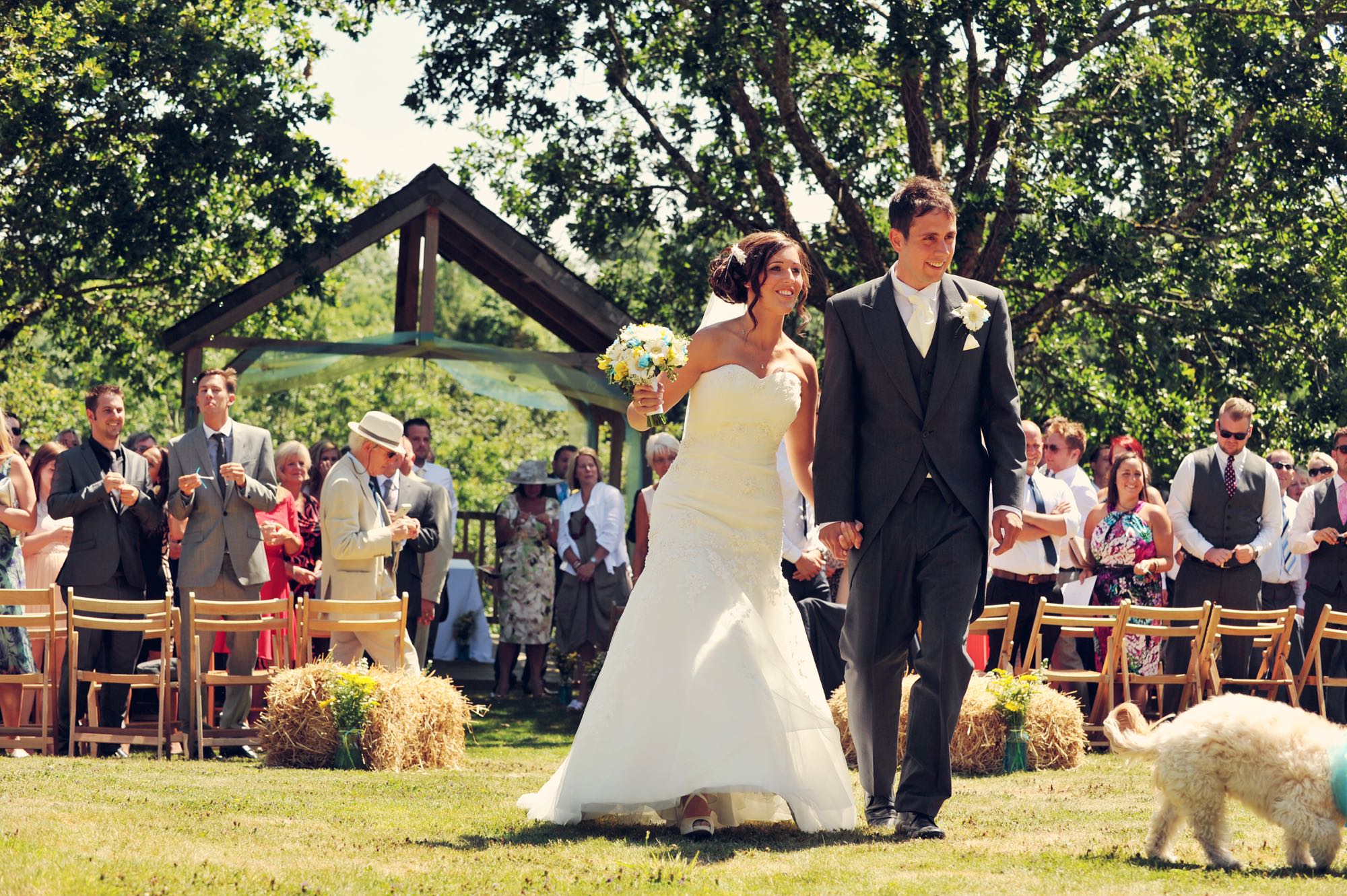 Newly weds leaving the Oak Arbour. The arbour is a wooden structure with four corner posts supporting a pitched roof. This photo shows a couple just walking away from the Oak Arbour after their ceremony. The arbour is decorated with a voile banner in tuquoise and yellow. The couple are in the middle of the shot, slightly offset to the right and the phot shows them in full length. They are holding hands and the groom is on the right, the bride on the left. Just slightly out of shot is the couples dog (you can't see his head). It is a summers day and the trees are in full leaf and the grass is parched. The bride is wearing an ivory, a-line, strapless dress. She has dark hair she is wearing up with a clip. In her right hand she is holding a bouquet of turquiose and yellow flowers. The groom is wearing a gray morning suit with white shirt tie. The couple are holding hands, smiling and looking forward and slightly to their left. You can see some guests in the background. The guests are looking at the couple and have their backs to the arbour. There are rows of chairs set out in front of the arbour with haybales at the end of the back rows. The guests are standing. You can seee the registrar standing by a clothed table under the arbour in the background.