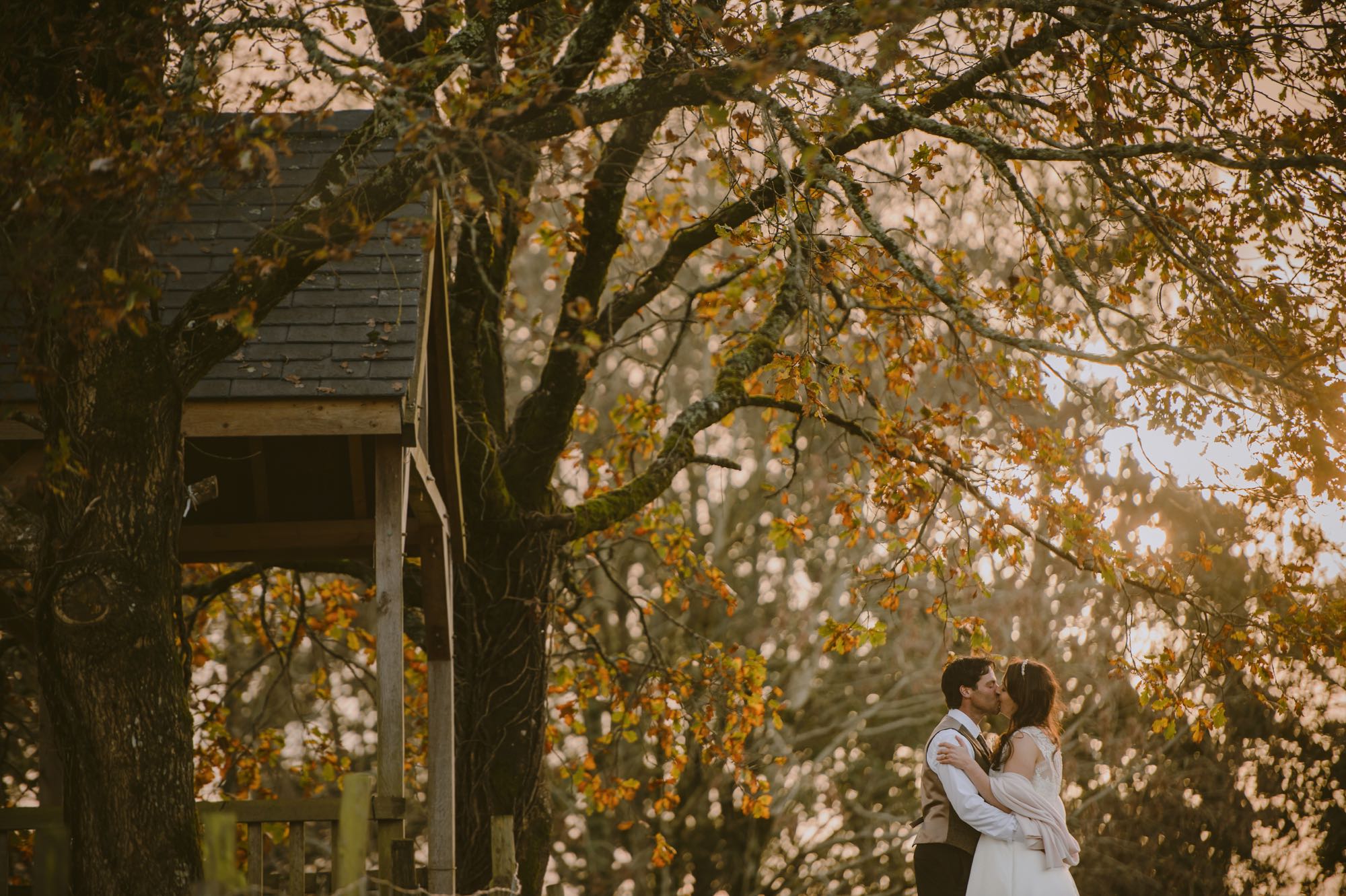 A couple having a romantic moment beside the Oak Arbour by themselves. This photo is taken side on to the arbour with the couple to the right of the shot. The arbour is a wooden structure with four corner posts supporting a pitched roof. The arbour has a large Oak Tree on each side. The trees are in autumn colours and the sun is shining through the branches. This picture is taken in the autumn. The couple are beside the arbour embracing. The couple are small compared to the trees and are shown from the knees up. The groom is on the left and the couple are embracing. The bride is wearing an ivory, short-sleeved, a-line dress. She has dark hair that she is wearing down. The groom is wearing a dark trousers a light bron waistcoat and has dark hair.