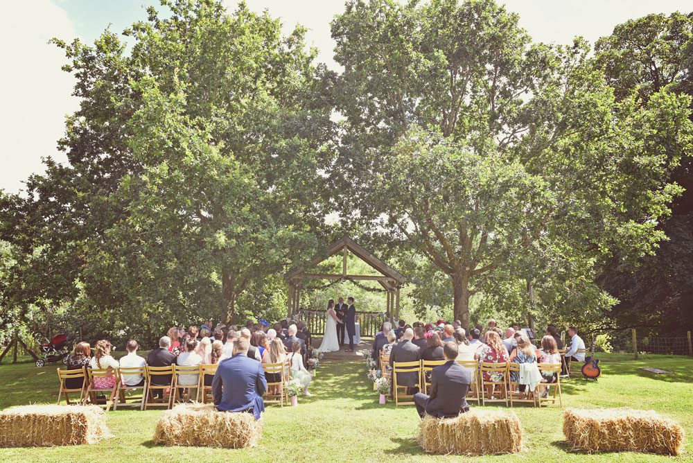 A couple exchanging vows under the Oak Arbour. The arbour is a wooden structure with four corner posts supporting a pitched roof. The arbour has a large Oak Tree on each side. The trees are in full leaf. This photo is taken from a bit of a distance and the whole of the Oak Trees are in the frame. The couple are under the arbour with the registrar and there is a clothed table (white) also under the arbour. The arbour is decorated with a garland of greenery. The couple are in the middle of the shot but are small compared to the canopy of the trees. They are holding hands and facing each other. The groom is on the right, the bride on the left. It is a summers day and the trees are in full leaf and the grass is a little parched. The bride is wearing a white, full length, sleeveless dress. She has dark hair she is wearing down. The groom is wearing a gray morning suit. The couple are holding hands and looking at each other. You can see there guests sitting in front of the arbour in two columns of rows of seats.. The guests are looking at the couple towards the arbour and have their backs to the camera. There is a row of haybales the back of the rows of chairs. You can seee the registrar standing by a clothed table under the arbour with the couple.