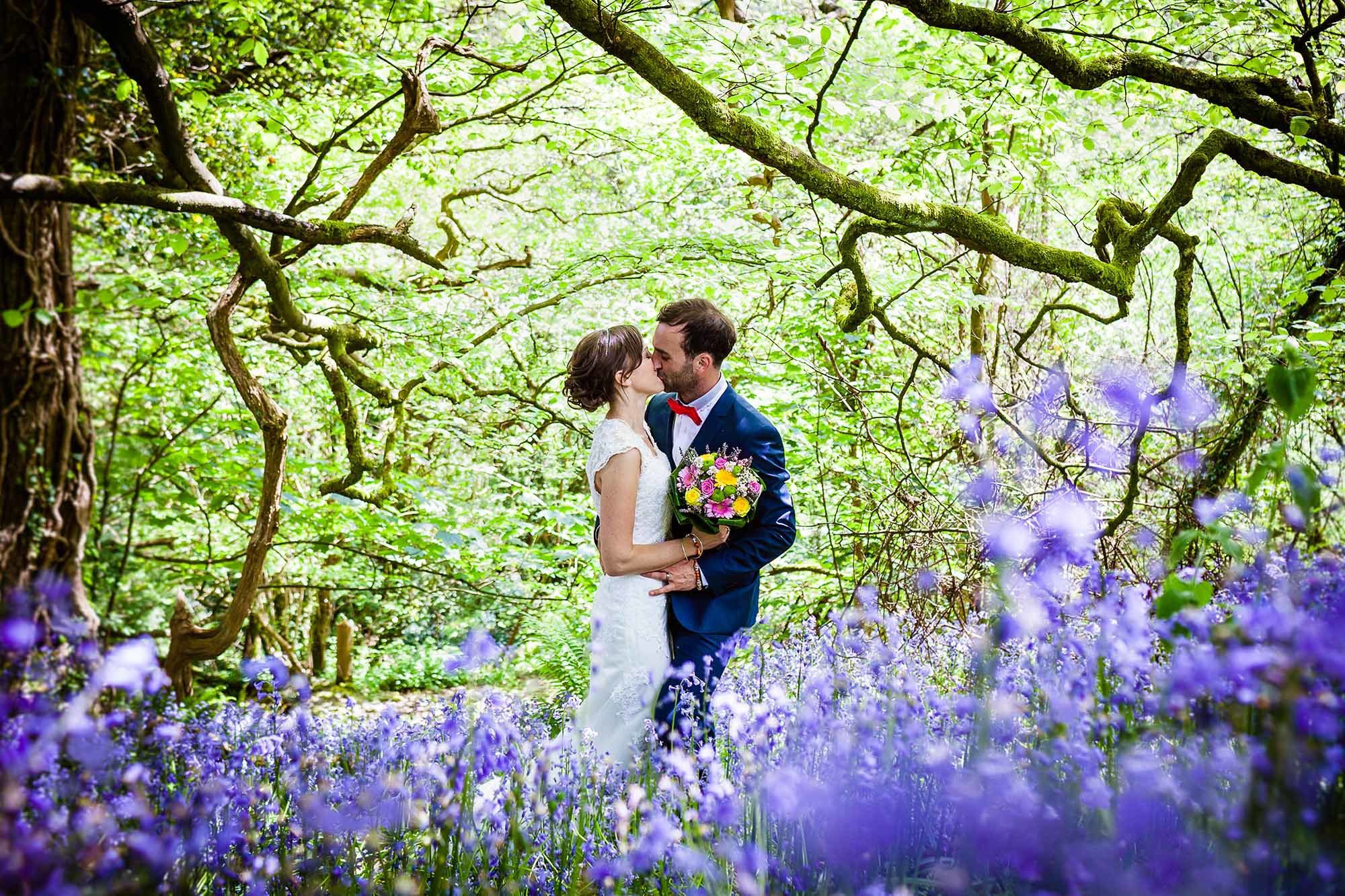 Newly weds kissing in a bluebell wood. The couple are in the centre of the image. They are surrounded by trees in leaf and there are lots of bluebells in the foreground. The image is taken in springtime. The couple are kissing the bride is on the left of the photo and the groom is on the right. The bride is wearing a lacy sleeveless dress you cannot see the couple's feet. In her right hand the bride is holding a bouquet of bright pink and bright yellow flowers. She has dark hair and she wears it up. The groom is wearing a mid blue suit with a white shirt and red bowtie. The groom has dark hair and a beard. The bride also has dark hair.