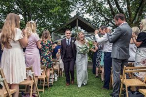 Newly weds leaving the Oak Arbour. The arbour is a wooden structure with four corner posts supporting a pitched roof. This photo shows a couple just walking away from the Oak Arbour after their ceremony. The arbour is decorated with a voile banner with stars on it. The couple are in the middle of the shot and the phot shows them in full length. They are holding hands and the groom is on the left, the bride on the right. The bride is wearing an ivory fitted dress with sparkles and lace details. It has cap sleeves. She has dark blond hair she is wearing loose. In her left hand she is holding a bouquet of green and cream flowers. The groom is wearing a dark brown 3-peice suit with gray shirt and a blue tie. The couple are holding hands, smiling and looking at each other. You can see some guests around them. The guests are looking at the couple and clapping and have their backs to the camera. There are rows of chairs set out in front of the arbour but the guests are standing.