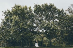 A couple having a romantic moment under the Oak Arbour by themselves. The arbour is a wooden structure with four corner posts supporting a pitched roof. The arbour has a large Oak Tree on each side. The trees are in full leaf. This photo is taken from a bit of a distance and the whole canopy of the Oak Trees is in the frame. The couple are under the arbour embracing. The couple are centred in the shot at the bottom of the frame. The couple are small compared to the canopy of the trees. They facing each other. The groom is on the right, the bride on the left. The trees are in full leaf and the sky is gray. The bride is wearing a white, full length, sleeveless, a-line dress. She has blond hair she is wearing up. The groom is wearing a navy jacket and chinos and has dark hair.