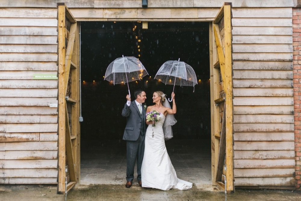 This image show s a couple standing in the doorway to the really rustic barn. The doors of the bar are open. The doors of the barn are very large the height is around twice the height of the couple. The doors open outwards. The couple stand just on the threshold and the barn looks dark on the inside. You can see two strands of festoon lighting inside the barn. The outside is clad in horizontal oak planks. The couple are holding their umbrellas above their heads. They are standing side-by-side, shoulder to shoulder facing the camera but looking at each other. The groom is on the left and the bride is on the right. The groom has dark hair and is wearing a gray three-piece suit with white shirt and white tie. The bride has blond hair which she is wearing up she has veil. The bride is wearing a fitted sleeveless full length white gown in silk. In her right hand she is carrying a bouquet with white flowers and greenery.