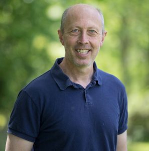 This is a head and shoulders shot of Mike. Mike works behind our bar and also looks after maintenance. He is wearing a blue polo shirt. The photo is taken outside in front of some trees. Make has gray hair and is smiling.