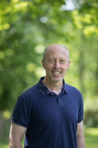 This is a head and shoulders shot of Mike. Mike works behind our bar and also looks after maintenance. He is wearing a blue polo shirt. The photo is taken outside in front of some trees. Make has gray hair and is smiling.