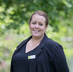This is an image of Charlie who is one of our events staff. This photo is a close-up shot of Charlie from the waist up. In the background there are green trees. Charlie is wearing a black top and a black blouse over this. She is also wearing a name badge. She has dark hair and is wearing it up and she is smiling.