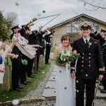 A couple walk away from the green room bar under the arch formed by swords. While they walk away their guests throw pom-pom confetti at them them. The bride is on the left and the groom is on the right. The groom where is a naval dress uniform. The bride wears a white, floor length gown with shoestring strap white and carries white and green flowers. The Green Room is a wooden chalet style building with glass frontage. This is an April wedding and the sky is overcast.