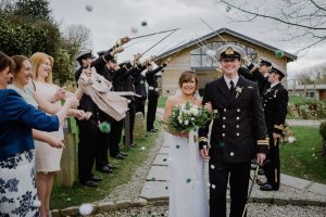 A couple walk away from the green room bar under the arch formed by swords. While they walk away their guests throw pom-pom confetti at them them. The bride is on the left and the groom is on the right. The groom where is a naval dress uniform. The bride wears a white, floor length gown with shoestring strap white and carries white and green flowers. The Green Room is a wooden chalet style building with glass frontage. This is an April wedding and the sky is overcast.
