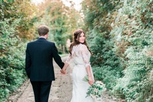 The bride and the groom are walking away from he camera down a narrow and leafy Cornish lane. The bride is on the right. She wears a fitted white gown with lace details and a delicate floral crown. The pic is takes from the back and the bride is looking over her right shoulder back at the camera. The groom wears a dark suit. The bride holds a bouquet of white and pink flowers in her right and it is hand-tied.