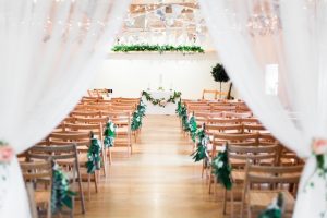 A shot of the wedding barn with oak floors and a-frame beams. There is greenery decorating the shelf at the back and also the edges of the 2 rows of folding wooden chairs. In the foreground there are 2 draped voiles with floral (pink rose) tie back.