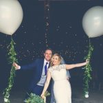 This is an image of the bride and groom standing in the doorway of the Really Rustic barn. They are standing side by side with the groom on the left. The bride is leaning slightly inwards towards the groom. The bride has a bouquet of greenery in her right hand. The couple each have their outside arms outstretched and each is holding a very large white balloon tethered by a "string" of greenery. The groom is wearing a navy suit, gray waistcoat, white shirt and blue tie. The bride wears a full-length, sleeveless, white dress with lace details and a beaded bodice.