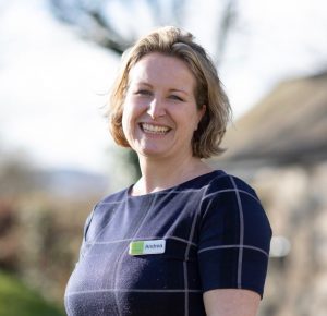 Andrea is standing outdoors at The Green. This shot is of her head and shoulders. Her left shoulder is slightly closer to the camera. She is smiling in the photo and is wearing a short sleeved, blue dress with a large chequered pattern. She hs chin length ash blond hair