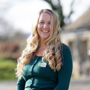 Cathryn is staning outdoors at The Green, She is standing slightly side on the the camera and this is a shot from the top of her head to her waist. She is smiling and has long, curly, blond hiar and is wearing a green too