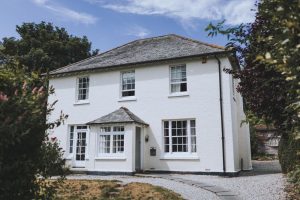 The House at The Green Cornwall showing a white front with three upper windows, two downstairs windows and entrance porch, blue sky and framed with greenery