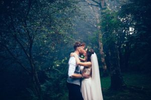Bridal couple embracing and kissing in a misty, woodland setting, the bride is wearing white with a long white veil fixed to the back of her hair and a natural garland headress, the groom has sunglasses on his head