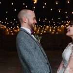 The couple standing in the doorway of the Really Rustic barn. The picture is taken from the waist up. Yoc an see festoon lighting in the background The are facing each other side on to the camera and holdinghands. The bride dark hair worn up. She has a fitted white dress with a sweatheart neckline and lace sleeves. The groom is wearing a tweed 3 piece suit and brown bow-tie.