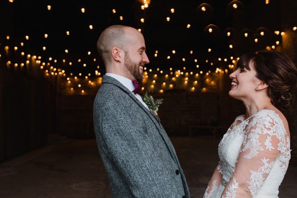 The couple standing in the doorway of the Really Rustic barn. The picture is taken from the waist up. Yoc an see festoon lighting in the background The are facing each other side on to the camera and holdinghands. The bride dark hair worn up. She has a fitted white dress with a sweatheart neckline and lace sleeves. The groom is wearing a tweed 3 piece suit and brown bow-tie.