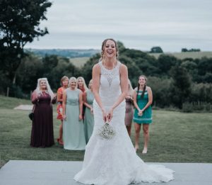 The bride is standing in the foreground facing the camera. She is wearing a white, sleeveless gown with a high neck and lace details. Behind her onthe lawn there is a group of girls standing on a lawn who are waiting for the bride to throw her bouquet. In the background you can see trees and rolling hills. The lawns are parched giving the feeling that it is the height of summer.