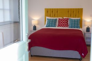 Open door leading to bedroom with Kingsize bed, red throw, yellow headboard and geometic design cushions, lamps on either side, Room 6 at The Green Cornwall