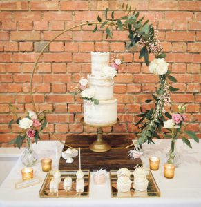 Three tier cake encircled with floral hoop and selection of cake treats, bonbons against a red brick background