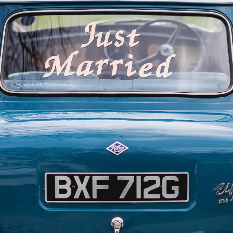 This image shows the rear of a Riley Elf car. It is a close-up shot of the back of the car and there is a saying in white writing on the rear window saying just married. Riley elf is blue in colour and the registration number is VXF712G.