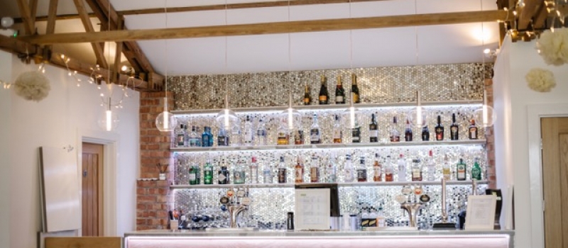 View of the Bar in the Red Brick Barn at The Green Cornwall with wooden front, three shelves of coloured bottles, sparkly tiled backdrop and beams above