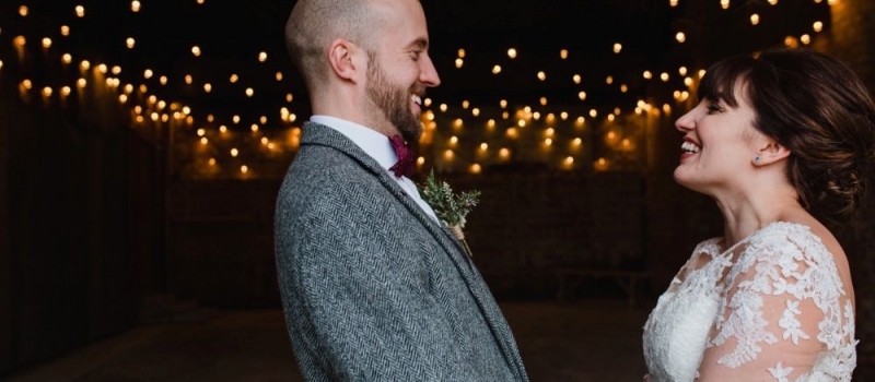 The couple standing in the doorway of the Really Rustic barn. The picture is taken from the waist up. Yoc an see festoon lighting in the background The are facing each other side on to the camera and holdinghands. The bride dark hair worn up. She has a fitted white dress with a sweatheart neckline and lace sleeves. The groom is wearing a tweed 3 piece suit and brown bow-tie.
